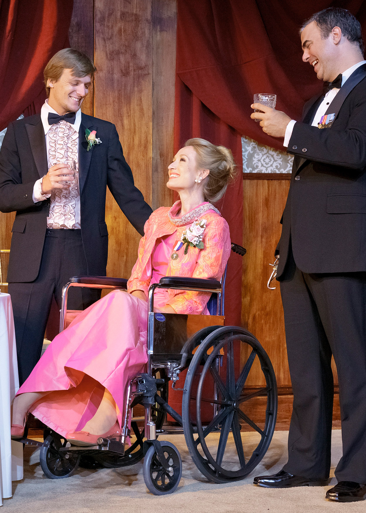 A woman in a pink dress sitting in a wheelchair between two standing men in tuxedos