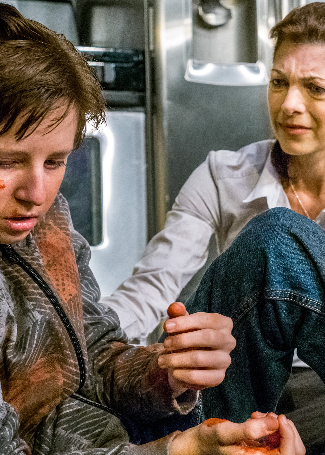 Production Photo from The Beautiful Dark (2013). A wounded boy sits on the floor of a kitchen and an older woman comforts him.