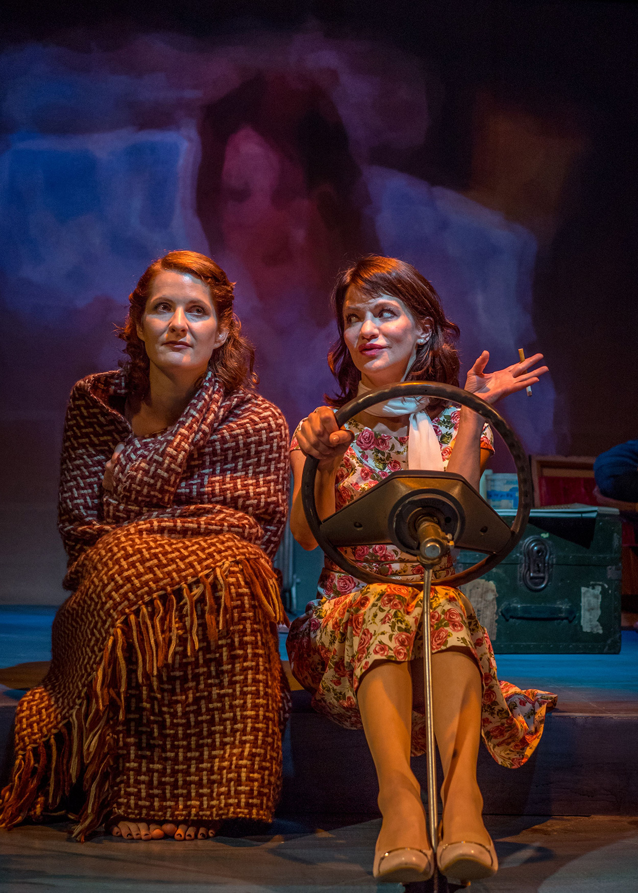 Production Photo from Janice Underwater (2014). Two women sit side-by-side. The woman to the left is wrapped in a blanket. The woman to the right is holding a car sterring wheels, depicting the women are in a car.