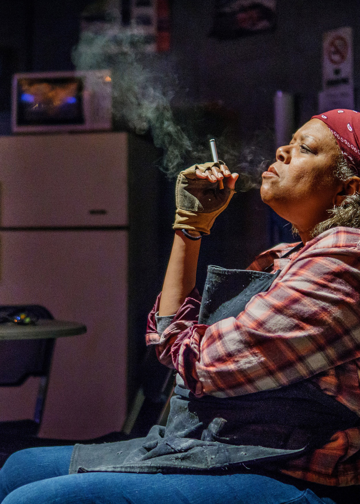Production Photo from Skeleton Crew (2017). A black woman sits in a dark room smoking a cigarette. She is wearing blue jeans, a gray apron, a red plaid shirt and a red bandana over her hair.