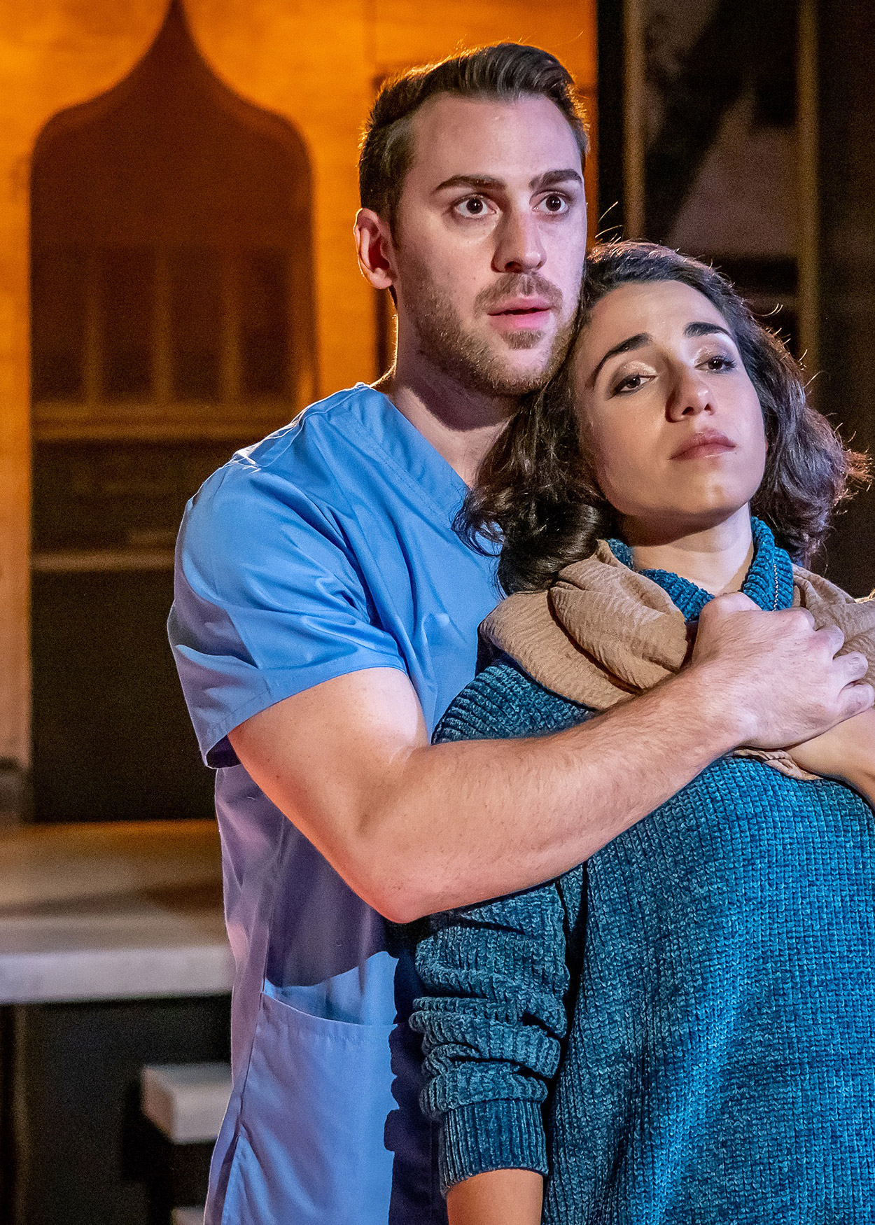 Production Photo from Yasmina's Necklace (2019). A man wearing a blue t-shirt stands behind a woman wearing a blue sweater, embracing her.
