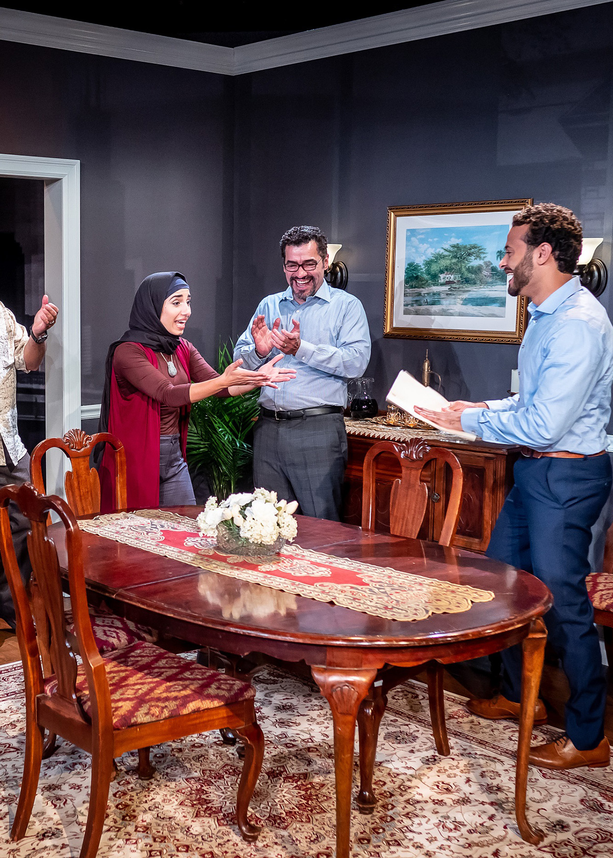 Production Photo from Yasmina's Necklace (2019). Four men and two women stand around a dining room table. Both women are wearing hijabs. One man is handing the younger woman a piece of paper. Everyone present is very excited.