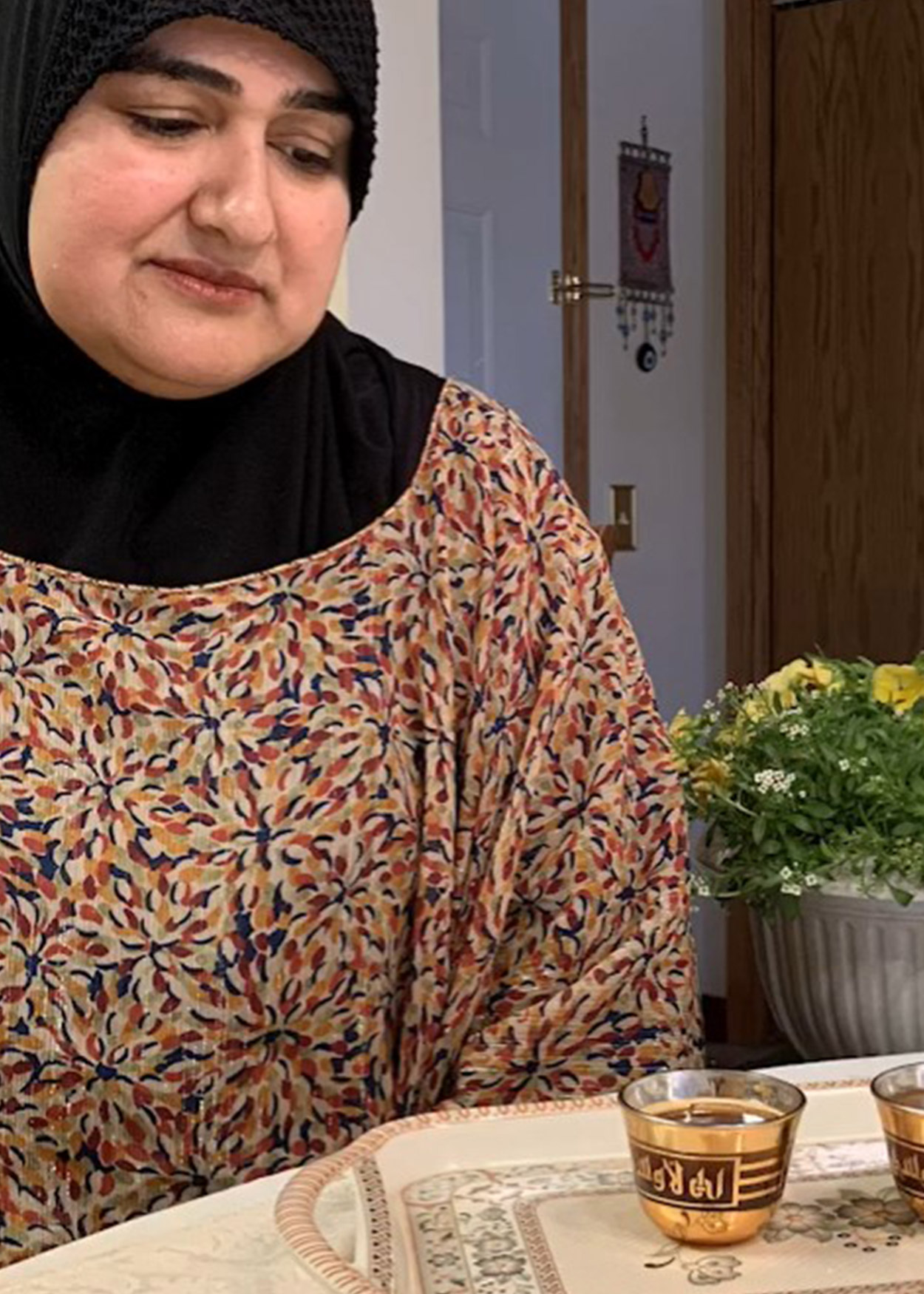 Image from Unveiled (2020). A South Asian woman, wearing a hijab, sitting at a kitchen table, looks at a cup of tea on a table.