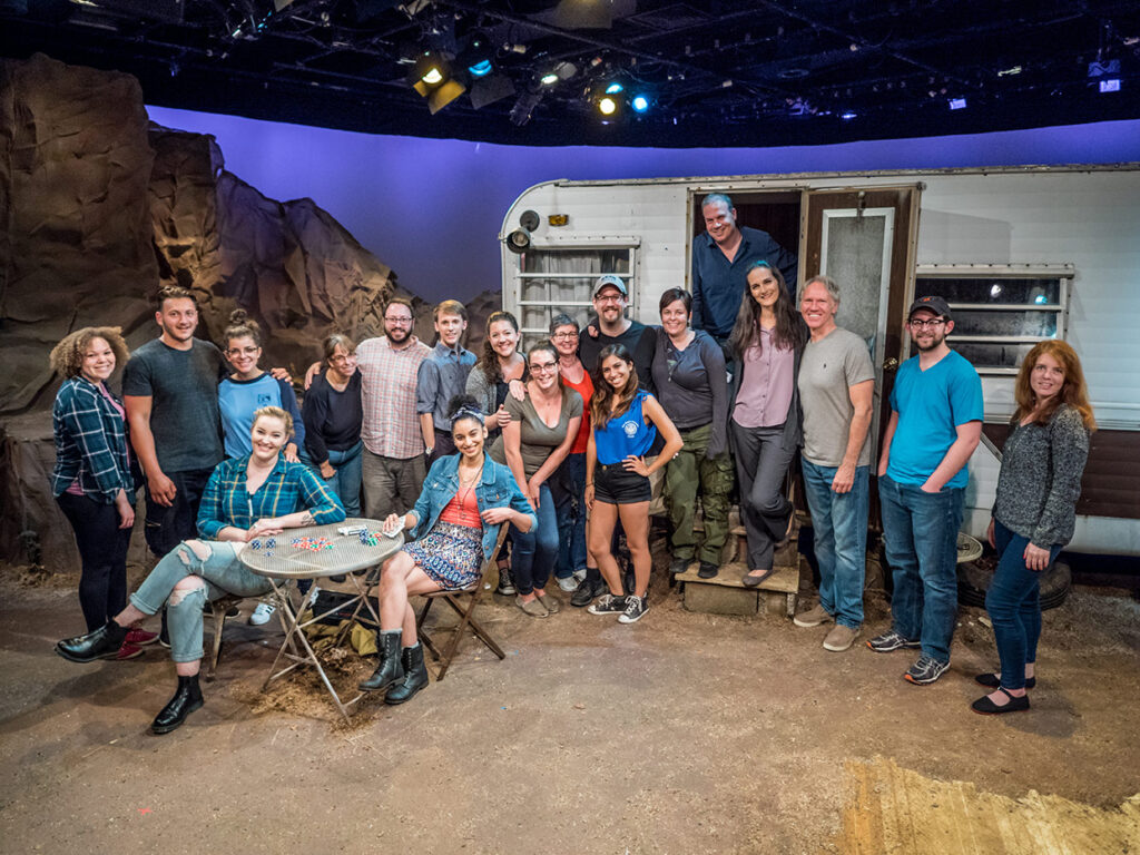 A production team of cast, crew, and interns gather on a set depicting a trailer in the desert