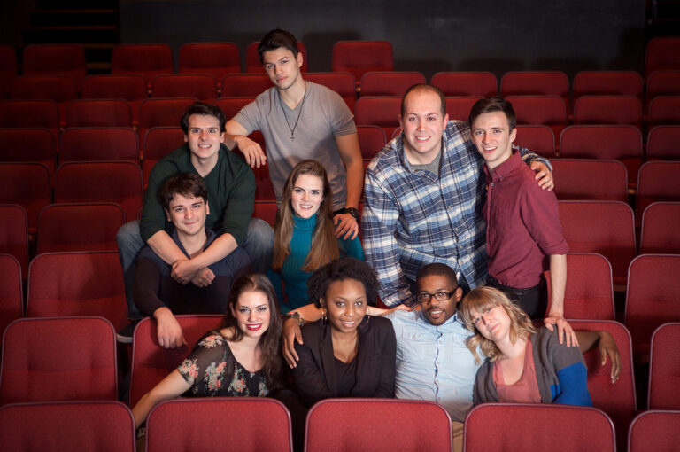 Group of young adults sit in a theatre amongst red chairs, posing for the camera