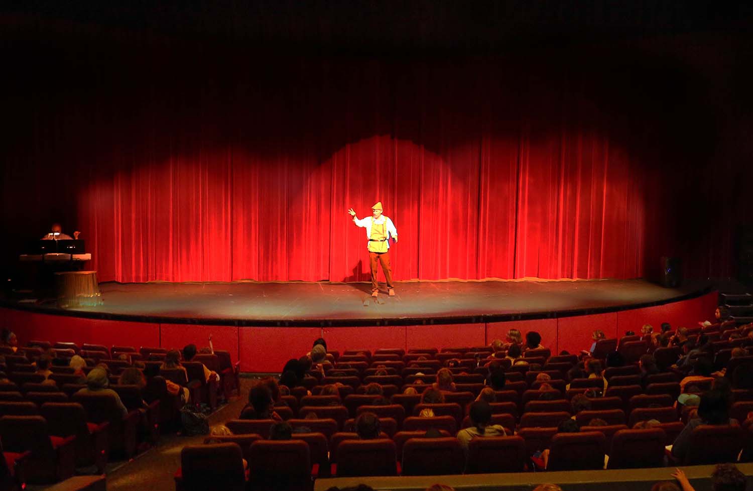 an actor on stage with red curtains behind him, speaking to a full house