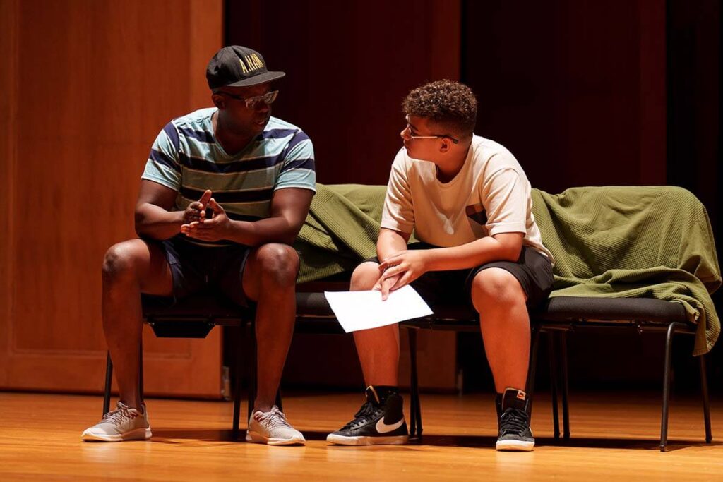 African American male sitting down and speaking to a student with curly hair, white shirt, Hispanic who holds what appears to be a play script and it's listening carefully.