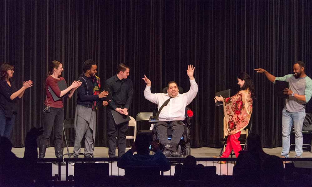 Actors on stage with the center actor in a wheelchair with a white shirt and his hands in the air, smiling