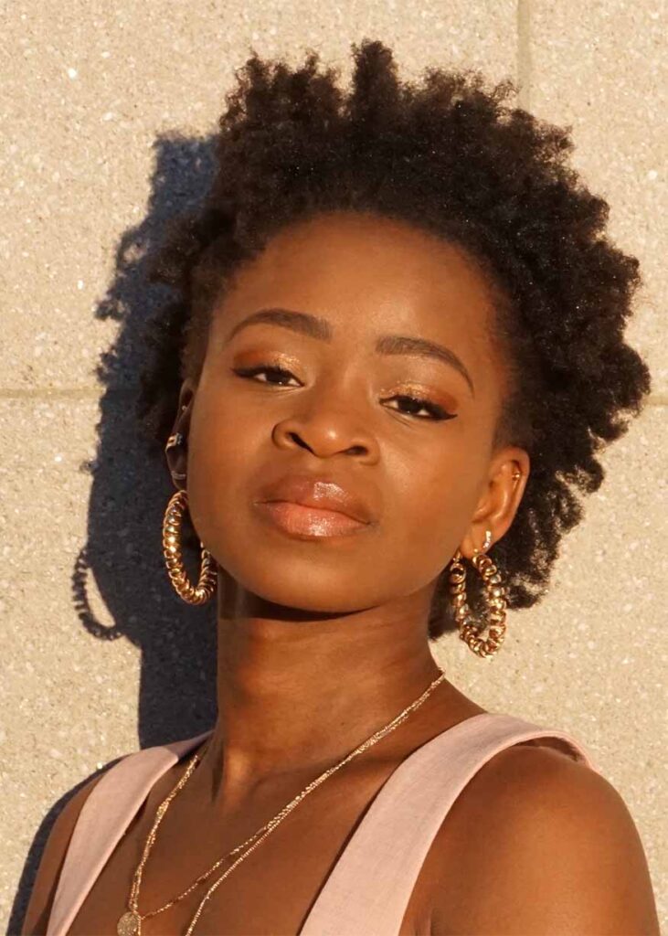 Playwright Gloria Majule is a young black woman. Her curly hair is cut into a cropped afro. She has gold make-up and is wearing gold hoop earrings and necklaces. She is leaning against a wall and is staring pensively into the camera. She is wearing a light pink tank top.