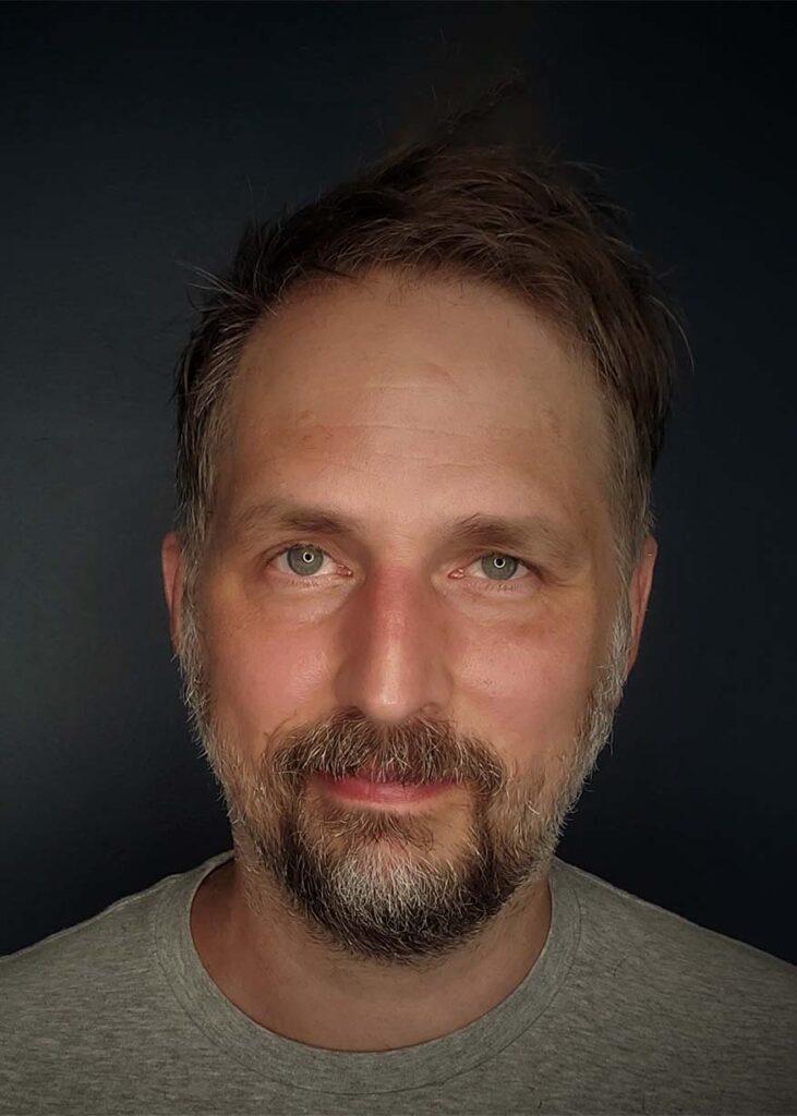 Playwright Scott Organ is a white man. His hair and beard have a mix of blonde and gray hair. He has green-gray eyes and is grinning at the camera.He is standing against a slate gray background and is wearing a light gray t-shirt.