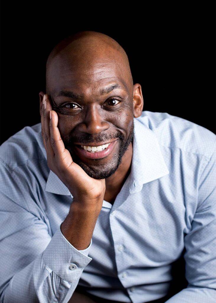 Playwright Vincent Terrell Durham is a black man. His head is shaven and has facial hair. He has a wide smile and his head is resting on the palm of his hand which is perched on a table. He is wearing a blue button up shirt.