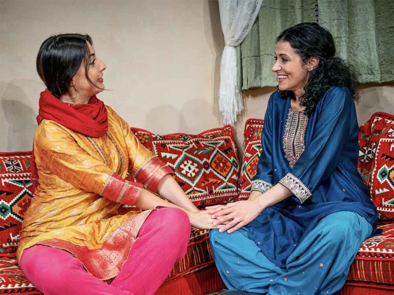 Two women sitting on a sofa, holding hands and smiling at each other as if they are close friends. they are dressed in colorful garments.