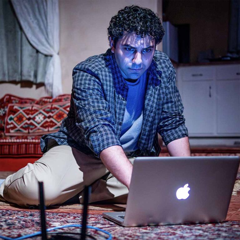 Actor Zaven Ovian stares at the glow emanating from a laptop in Premiere Stages’ 2023 production of Selling Kabul by Sylvia Khoury.