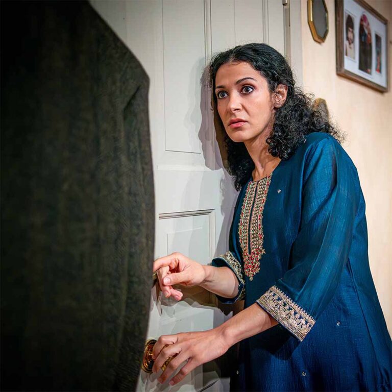 Actress Atra Asdou next to a door with a scared face, holding the nob, in wait.