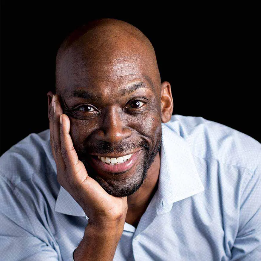 Playwright Vincent Terrell Durham is a black man. His head is shaven and has facial hair. He has a wide smile and his head is resting on the palm of his hand which is perched on a table. He is wearing a blue button up shirt.