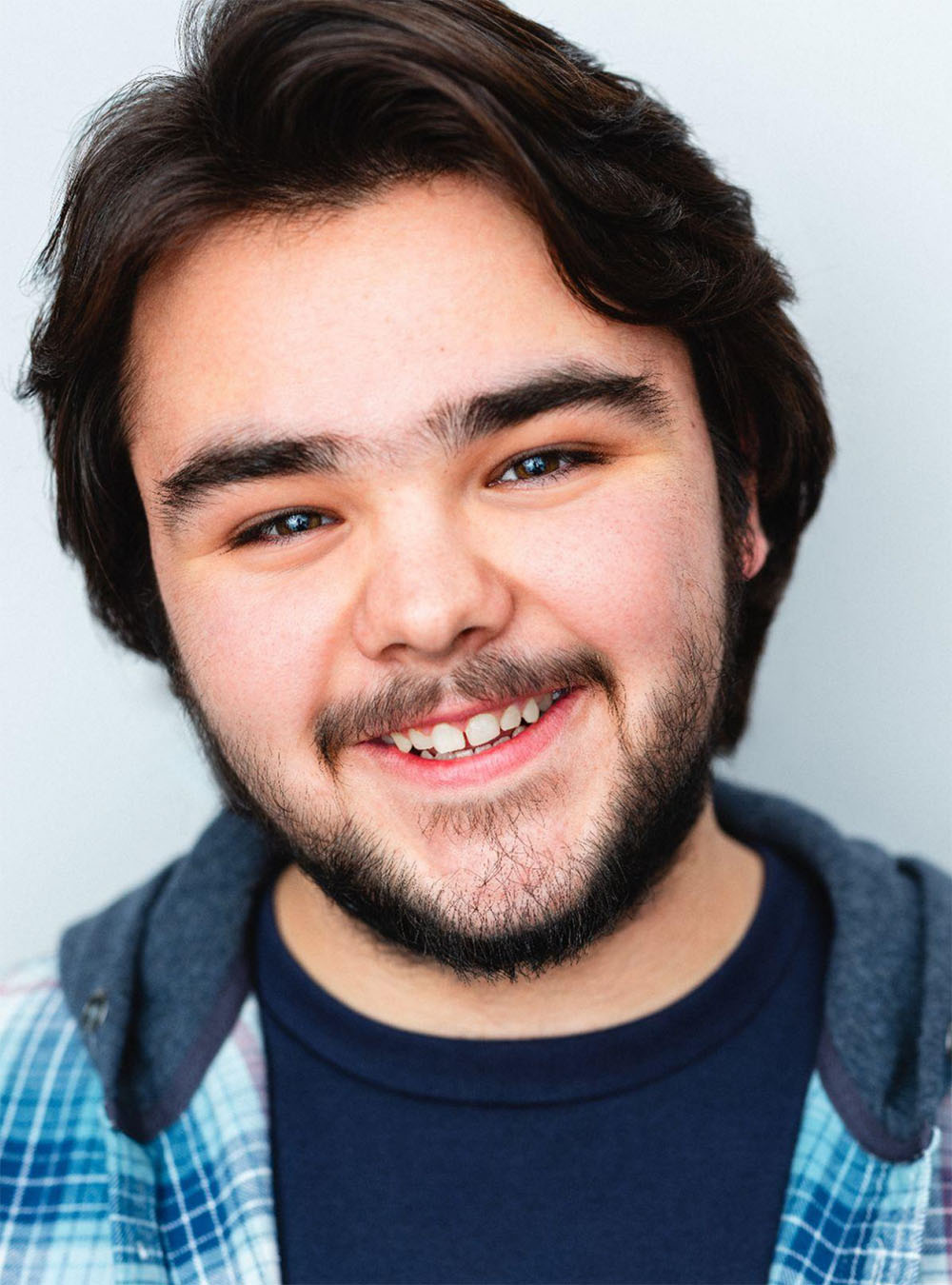 A cheerful young man with a friendly expression smiles warmly at the camera. He has dark brown hair and a neatly trimmed beard, wearing a casual blue plaid shirt over a navy t-shirt. The background is a soft, neutral tone, emphasizing the subject's approachable and joyous demeanor.