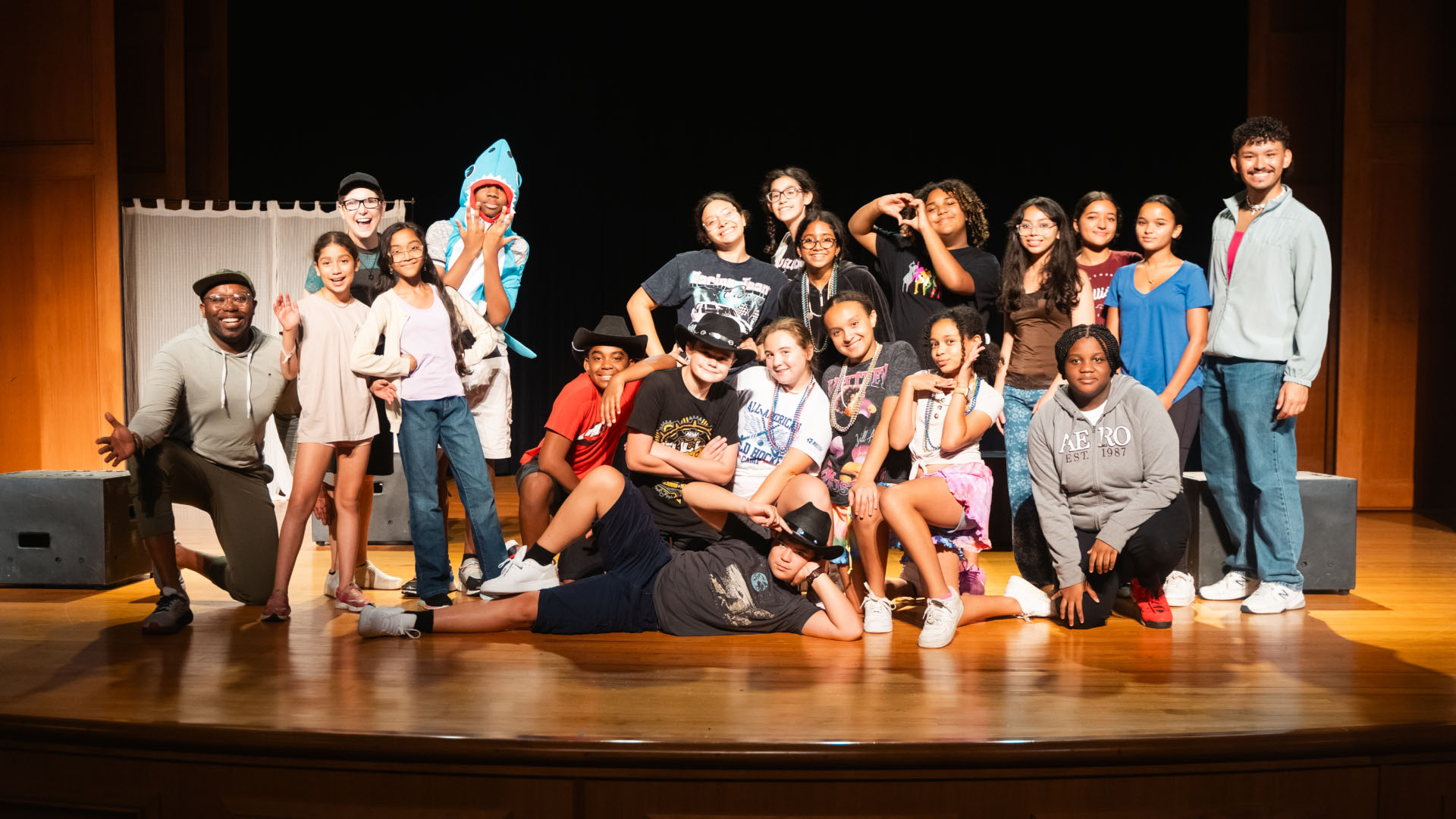 A large group of children and teens pose together on a brightly lit theater stage, arranged in playful and dynamic stances. Some participants are standing while others kneel or lie across the wooden floor, creating a lively, energetic group formation. A performer in a shark costume adds a humorous touch on the left side. The backdrop is simple and dark, drawing focus to the group and the warm stage lighting. The scene suggests a summer theater camp, drama workshop, or youth performance celebration.