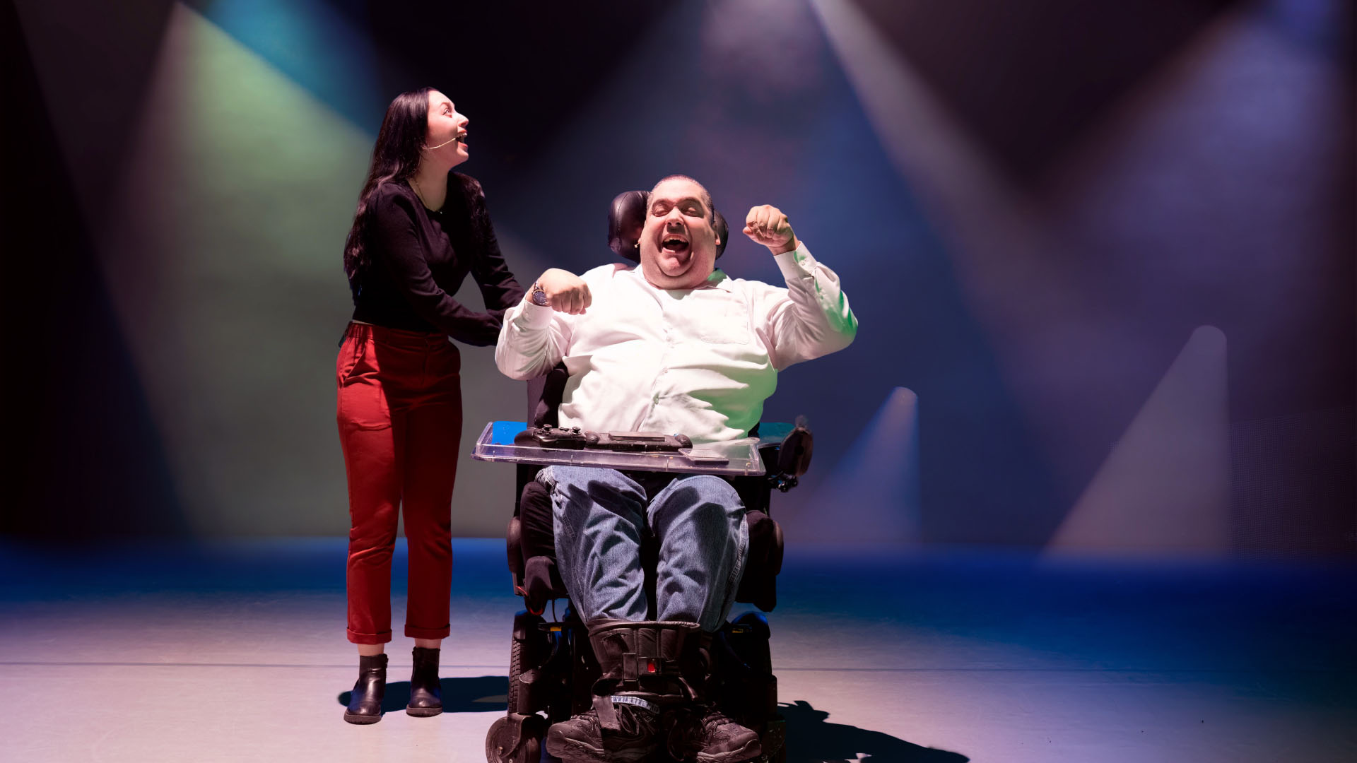 A man in a wheelchair and a woman share a joyful moment under colorful stage lights. The man, smiling broadly, is wearing casual attire and appears to be celebrating. The woman, standing beside him, looks upwards with a smile, wearing a black top and red pants.