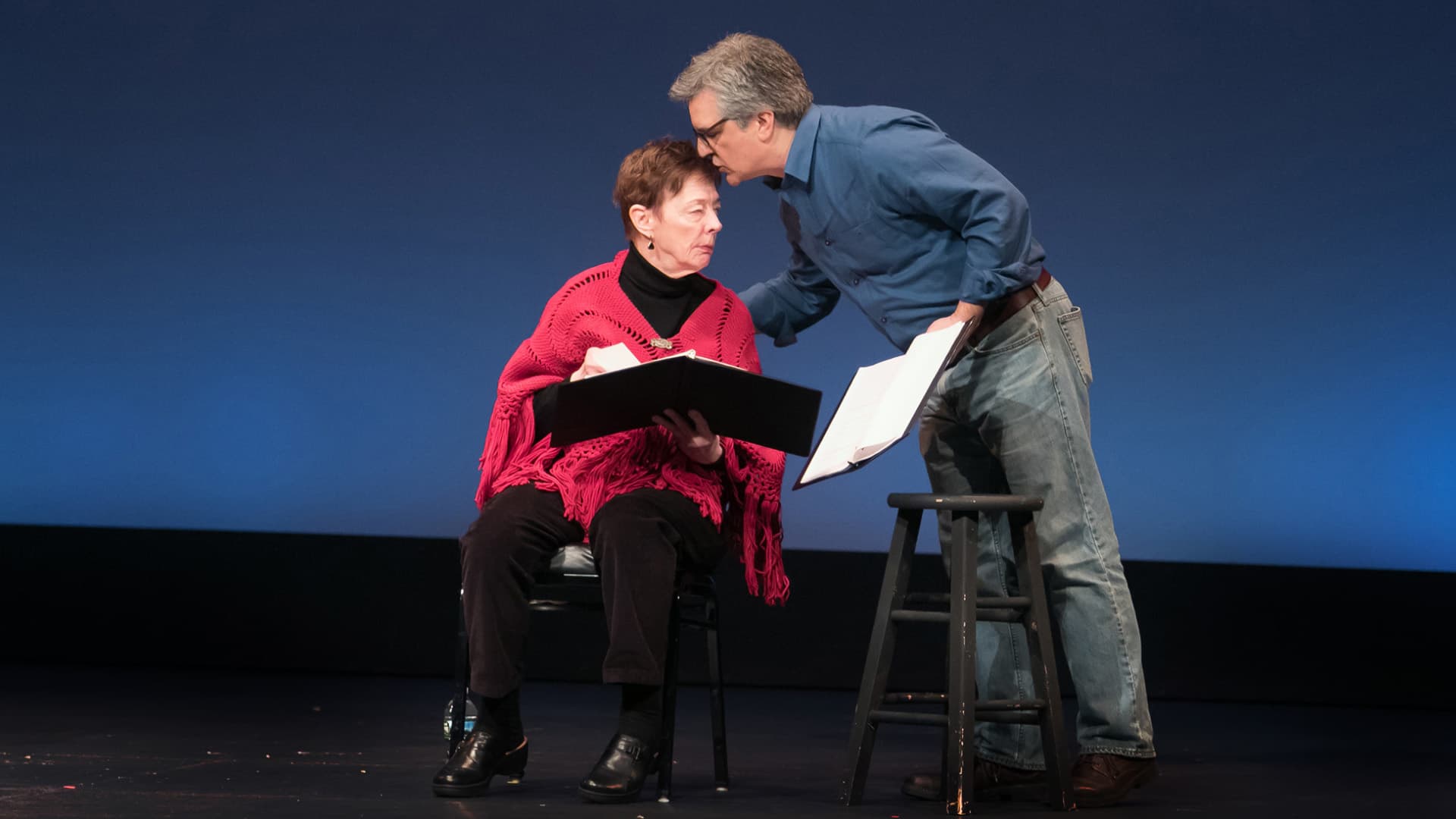 Two actors engage in a theatrical rehearsal, seated on stage with scripts in hand. The intimate setting emphasizes the collaborative nature of drama and the importance of dialogue and expression in live theater, showcasing the creative process of character development and scene preparation.