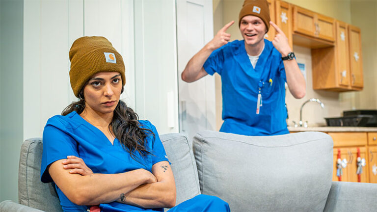 A female actor in blue scrubs sits on a couch, looking frustrated or annoyed, with her arms crossed. A male actor, also in blue scrubs, stands behind her, wearing a brown beanie and making playful gestures as if teasing her.