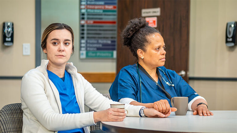 Two female actors in a hospital break room setting. They are seated at a table with coffee cups in front of them. Both are dressed in medical scrubs, with the woman on the right appearing to be older and looking attentively at something off-camera, while the younger woman on the left gazes forward, expressionless.