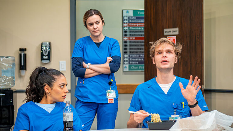 Three healthcare workers in blue scrubs are seated at a table in a hospital break room. The nurse in the middle stands with her arms crossed, looking down at the nurse sitting on her left. The male nurse on her right gestures while holding a fork, mid-conversation.