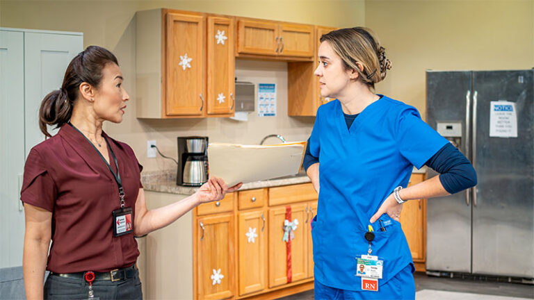 A nurse in a maroon shirt holds a file while speaking to another nurse in blue scrubs, who has her hands on her hips. They are standing in a break room with wooden cabinets, a coffee maker, and a refrigerator in the background.