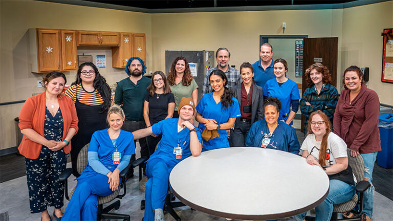 A diverse group of actors and crew members standing together on a stage set that resembles a break room in a hospital. Some of the cast members are wearing scrubs, while others are in casual clothing, indicating the crew behind the production. The room features a table, chairs, and kitchen cabinets in the background.