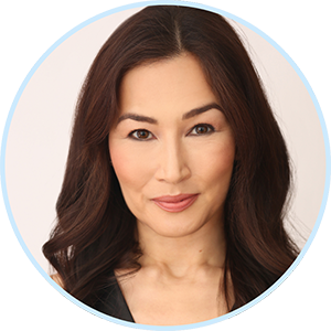 A woman with long, dark brown hair and light makeup smiles softly at the camera. She is wearing a black top and is posed against a plain, light background, framed within a circular border.