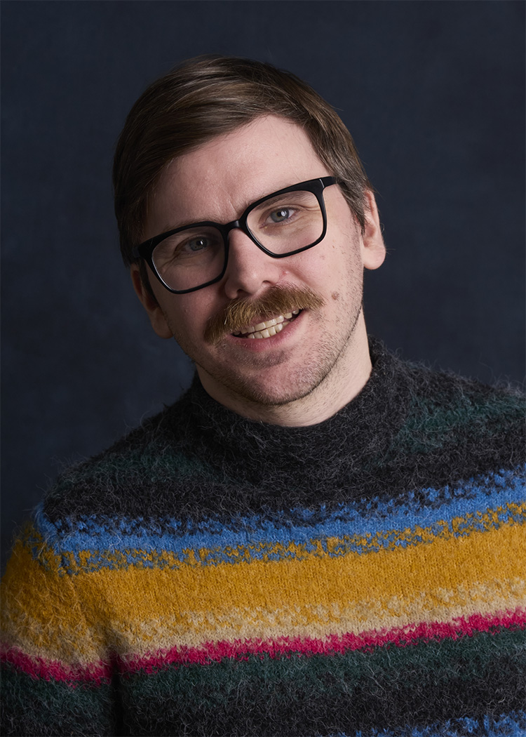 A man with glasses and a mustache smiles warmly against a dark background. He wears a fluffy, striped sweater featuring vibrant colors like yellow, blue, and pink, adding a cheerful and casual vibe to the portrait. The lighting and focus emphasize a friendly and approachable demeanor.