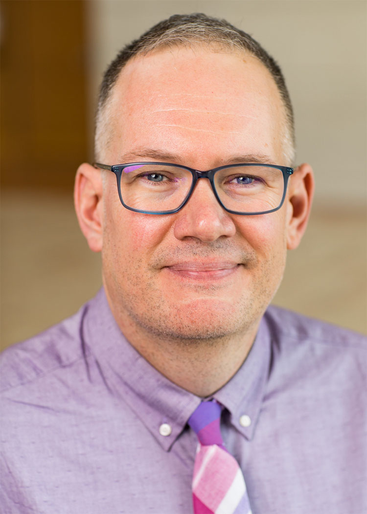 Erik is a middle-aged white male with short, cropped hair. His gray-blue framed glasses match his eyes. He is wearing a purple button up shirt and matching plaid purple tie. He has a tight lipped smile.