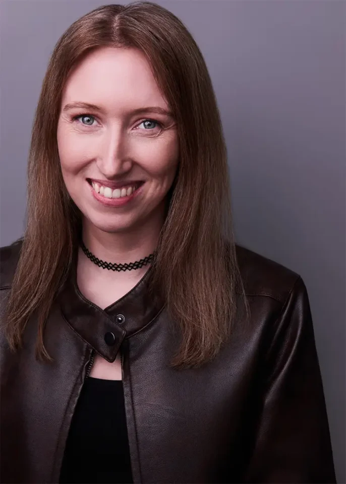 A stylish woman with long brown hair smiles warmly, wearing a dark leather jacket and a choker necklace against a neutral gray background.