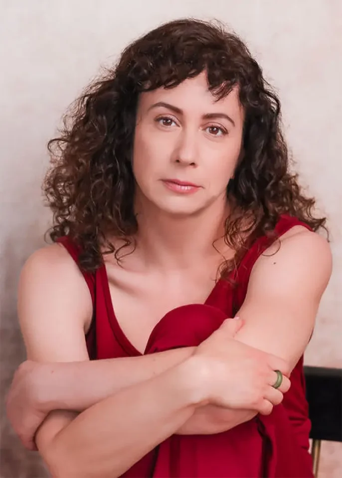 A woman with curly brown hair sits pensively, wearing a red dress. Her expression is introspective against a beige background.