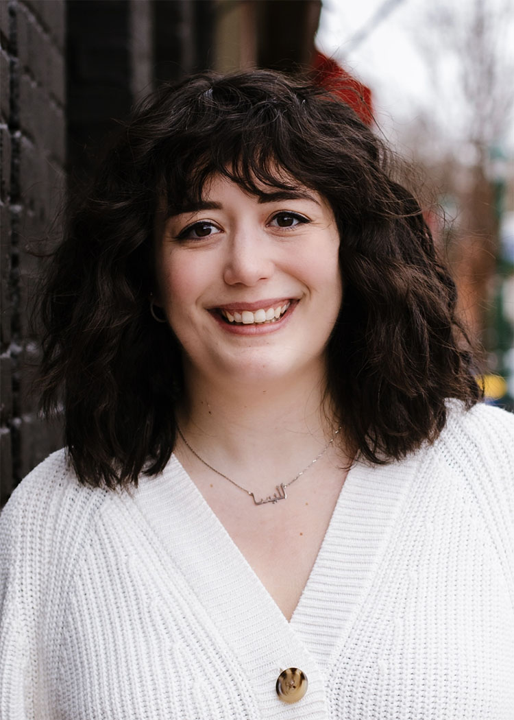Alyssa is an Arab-American playwright. She has dark, shoulder length curly hair, brown eyes and is wearing a white button up sweater. She has a bright, welcoming smile.