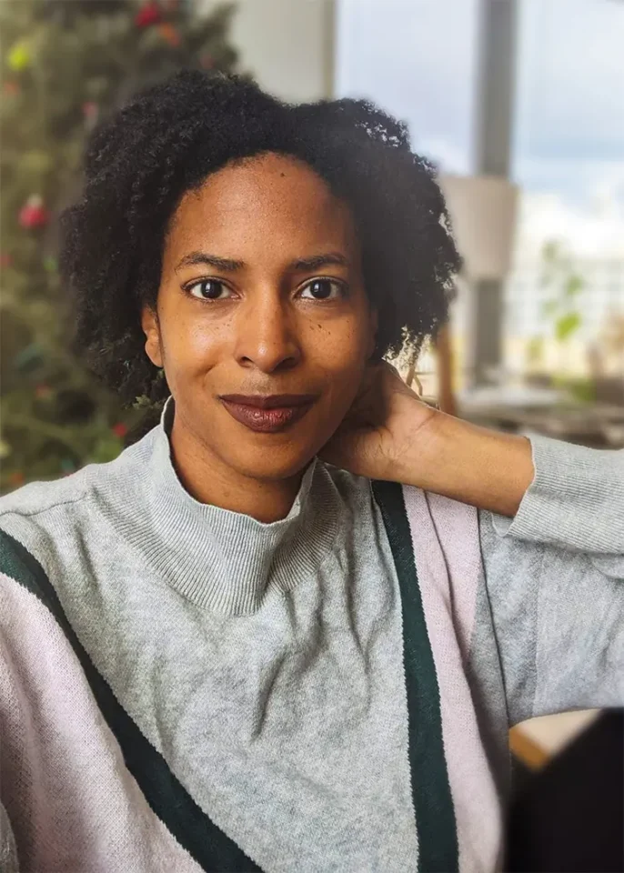 A woman with an inviting smile poses indoors with a stylish sweater, set against a soft-focus backdrop featuring a decorated Christmas tree and warm interior lighting.