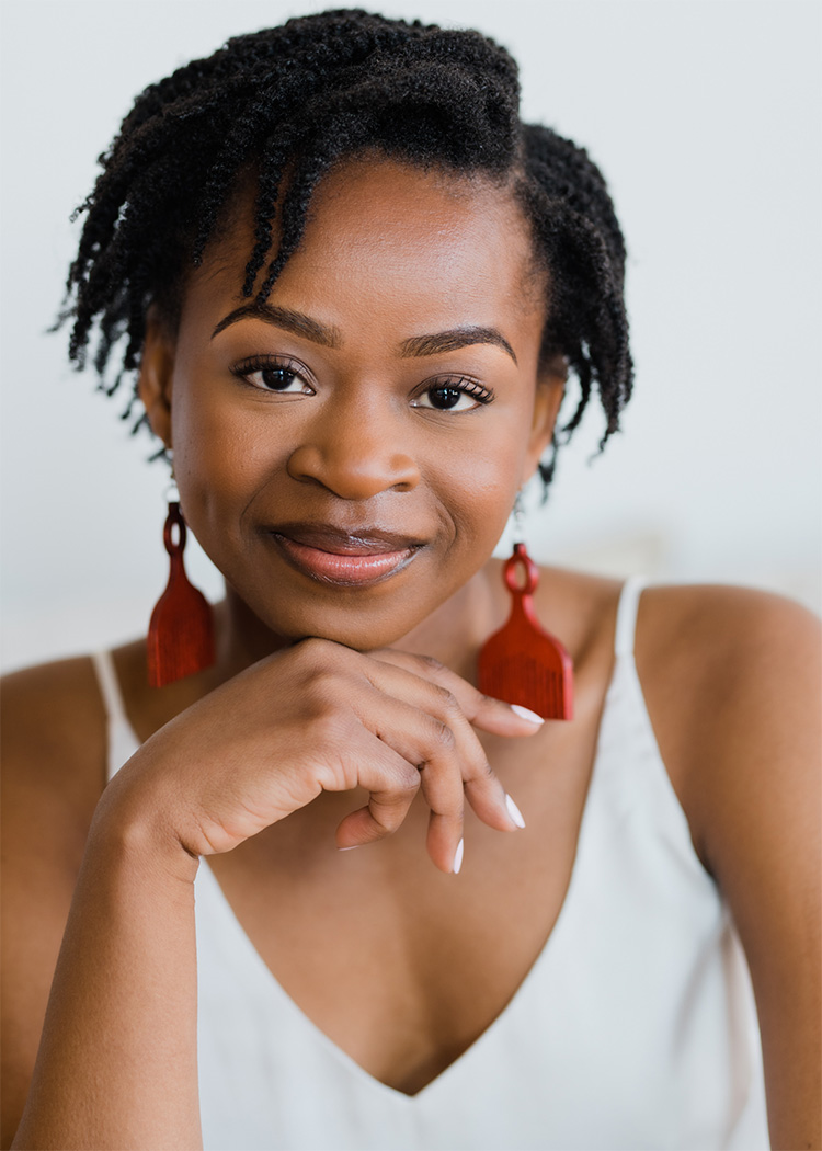 A close-up portrait of a woman smiling gently while resting her chin on her hand. She has short, curly hair styled naturally and wears striking red earrings contrasting with her light-colored outfit. Her expression exudes confidence and warmth.