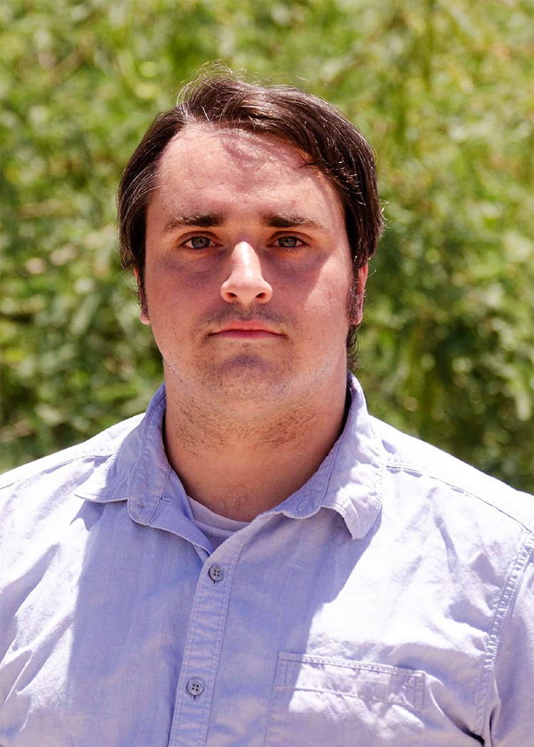 A man with short hair wears a light-colored button-up shirt while standing outdoors. The background is blurred greenery, emphasizing a natural setting. His expression is neutral, suggesting a candid or thoughtful moment.