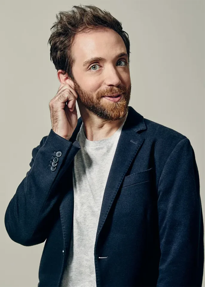 A man with a well-groomed beard wearing a casual dark blue blazer over a gray shirt, showcasing a relaxed yet confident demeanor. His hand is raised to his ear, suggesting attentiveness or thoughtfulness.