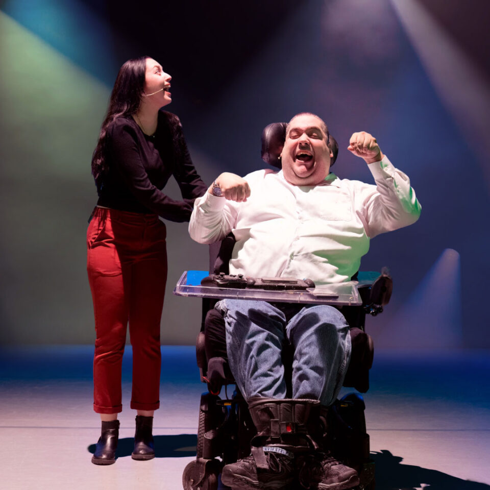 A man in a wheelchair and a woman share a joyful moment under colorful stage lights. The man, smiling broadly, is wearing casual attire and appears to be celebrating. The woman, standing beside him, looks upwards with a smile, wearing a black top and red pants.