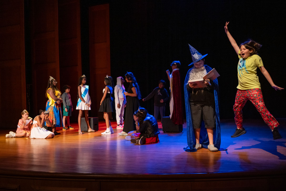 A group of children and teens perform on a theater stage dressed in colorful costumes, portraying characters such as a wizard, dancers, and fantasy figures. Some performers stand in a line at center stage, others sit or kneel in dramatic poses, while one child leaps energetically on the right side under a spotlight. The stage is warmly lit, with a dark backdrop that highlights the actors and their expressive movements, capturing the feel of a youth theater production or drama camp showcase.