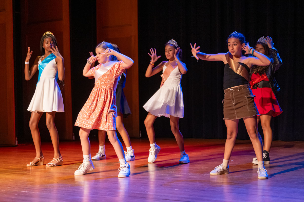 A group of young dancers energetically performs on a stage, each exhibiting individual style and flair. Dressed in a variety of colorful outfits, from casual to elegant, they engage the audience with synchronized movements and expressive gestures.