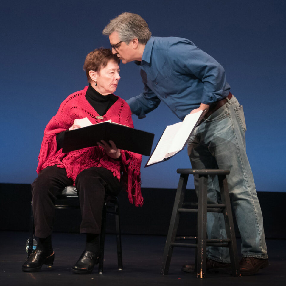 Two actors engage in a rehearsal on stage, featuring a seated woman in a red shawl and a man holding a script, leaning in for close interaction. The setting emphasizes the intimate nature of the theatrical process, with simple props and a focused interaction