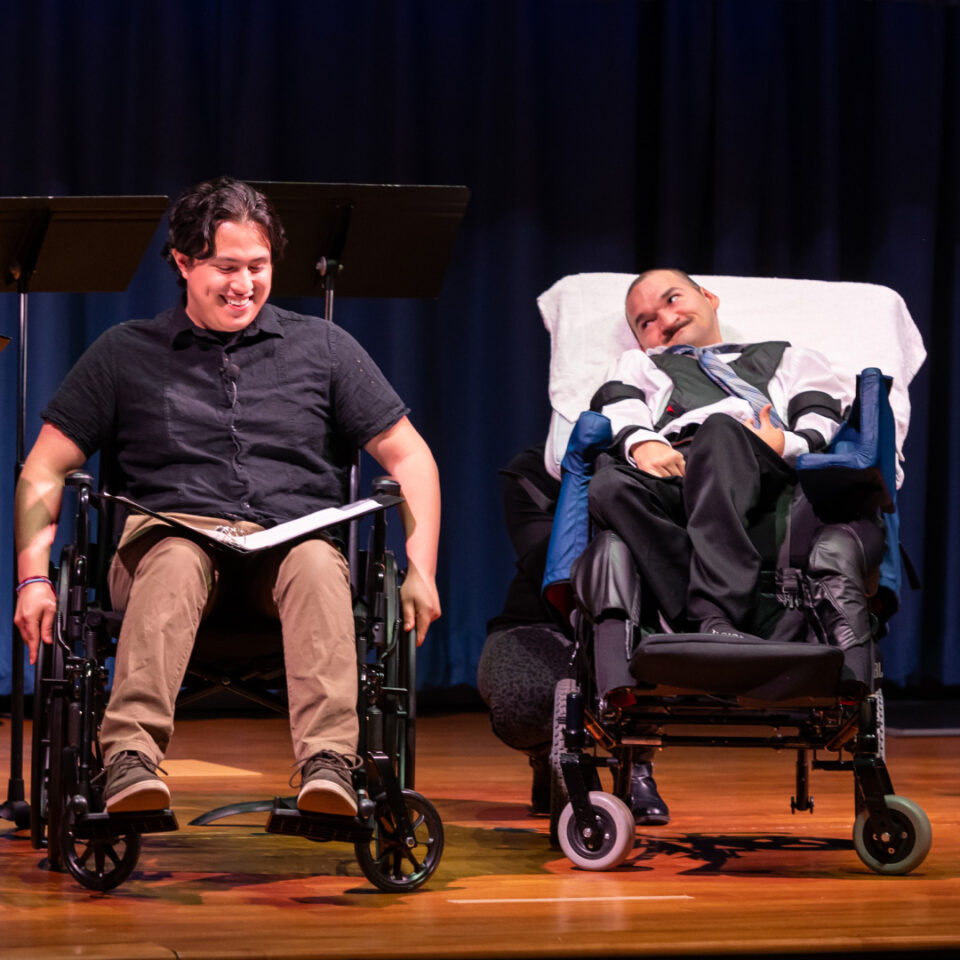 Two men are sitting in wheelchairs on a stage, engaged in a conversation or possibly a performance. Both individuals appear to be smiling, creating a warm and positive atmosphere. The setting suggests an inclusive event, emphasizing accessibility and community involvement. The stage is softly lit, focusing attention on the two individuals as main subjects.