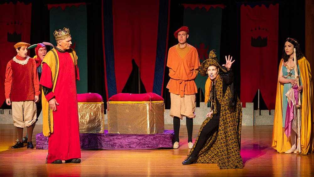 A set of actors dressed up in medieval wardrobes on stage, with a witch-like character.