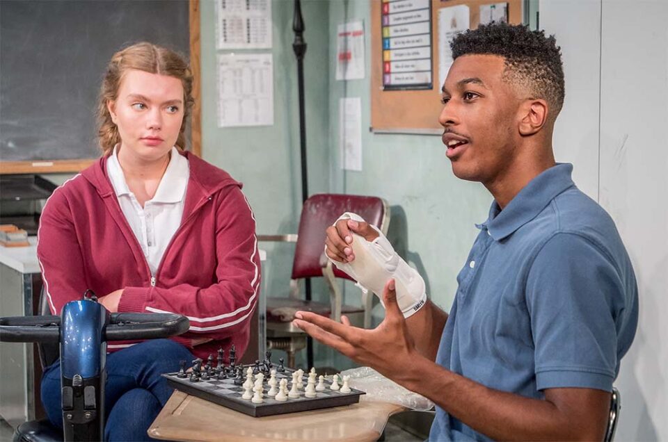 Two people sitting at a table with a chess board on it. One is a woman wearing a red hoody, and a man, with a hurt hand, speaking, wearing a blue shirt.