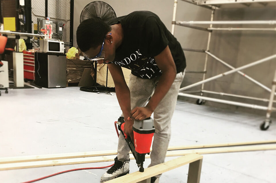 Student wearing a black shirt and jeans, using a building tool, stapling wood parts.
