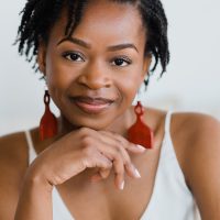 A close-up portrait of a woman smiling gently while resting her chin on her hand. She has short, curly hair styled naturally and wears striking red earrings contrasting with her light-colored outfit. Her expression exudes confidence and warmth.