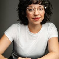 Playwright A woman with curly hair wearing glasses smiles gently while seated. Her outfit is casual, featuring a plain white t-shirt. The background is neutral, emphasizing her relaxed and approachable demeanor. The image captures a sense of warmth and friendliness, highlighted by her soft expression and natural pose.
