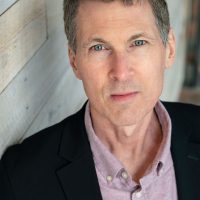 PLAYWRIGHT A man with short brown hair and blue eyes is captured in a professional headshot. He wears a black blazer over a light pink button-up shirt. His expression is neutral and he was photographed against a background of light wooden panels, adding texture and warmth to the scene.