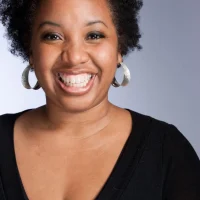 A woman with curly hair wearing hoop earrings smiles warmly. Her black top contrasts against a neutral background, creating a lively and engaging portrait.